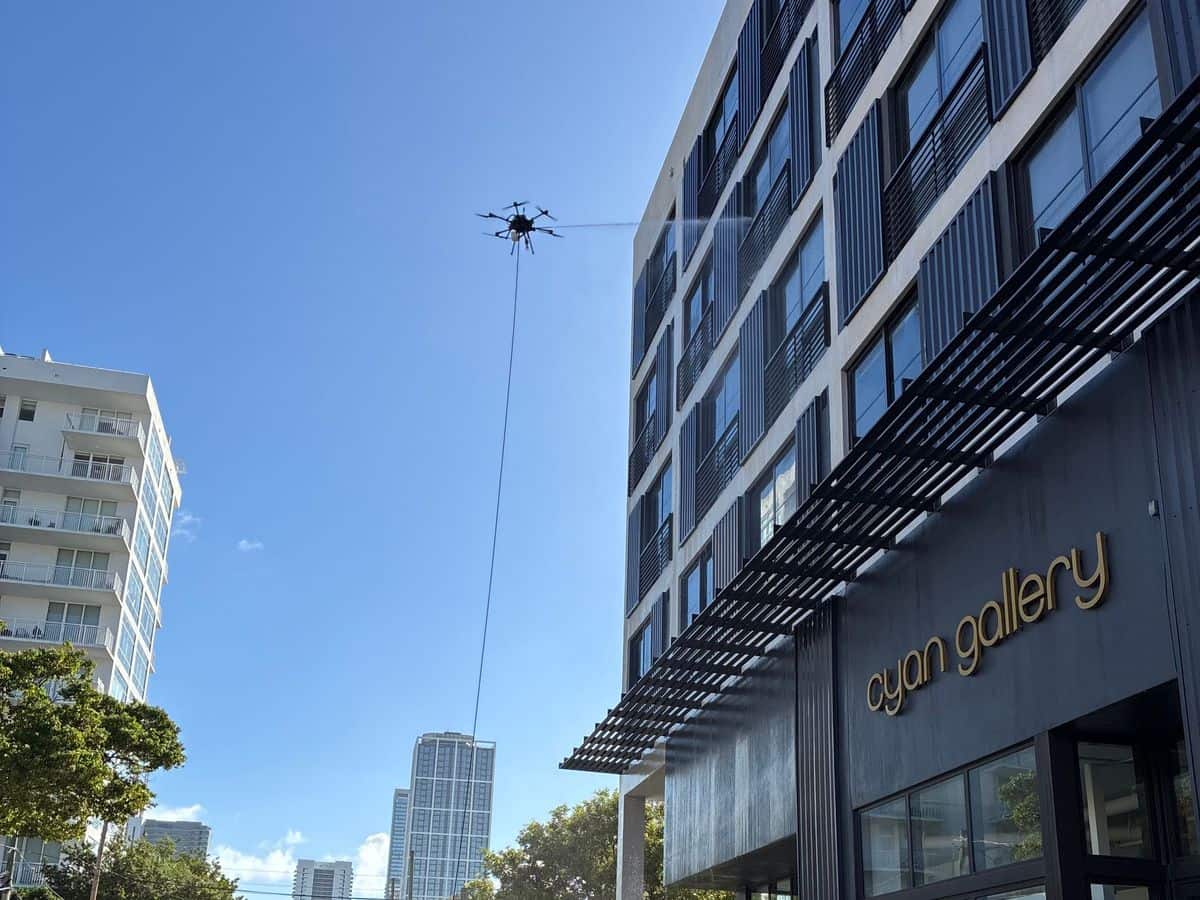 The tethered cleaning drone washing the upper windows of Sentral Wynwood above a street-level gallery, with cones and the hose along the curb